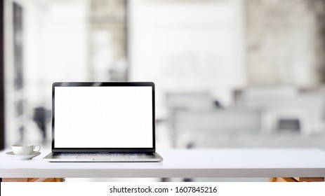Cropped shot of simple workspace with blank screen laptop and coffee cup on white table with blurred office room background
