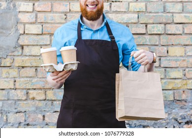 Cropped Shot Of Smiling Male Barista In Apron Holding Paper Bags And Disposable Cups With Coffee