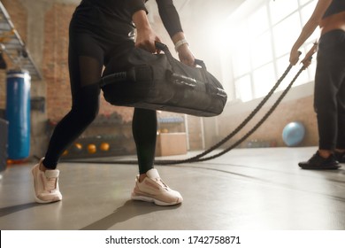 Cropped shot of sportive woman lifting a heavy sandbag while having workout at industrial gym. Group training, teamwork concept. Selective focus