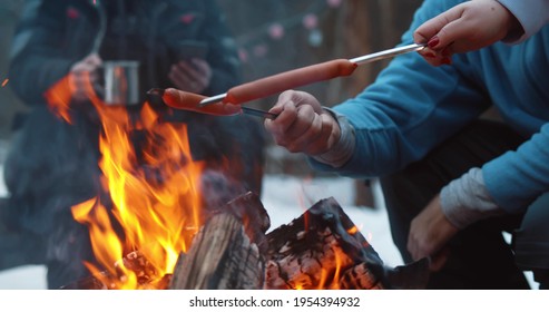 Cropped Shot Of Travelers Baking Delicious Sausages On Campfire In Winter Forest. Group Of Friends Hiking And Camping In Winter Woods And Preparing Food On Fire