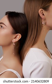 Cropped shot: two ladies are showing creative asymmetric earrings made as enamelled metal flowers with thin chains, crystals and pearls. Ladies are standing next to each other on the gray background. 