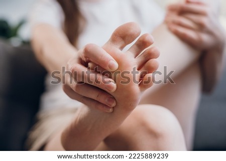 Cropped shot of unhealthy young female massaging her bunion toes to relieve pain sitting on grey couch at home. Sick woman holding painful feet. Toes pain, arthritis and healthy concept