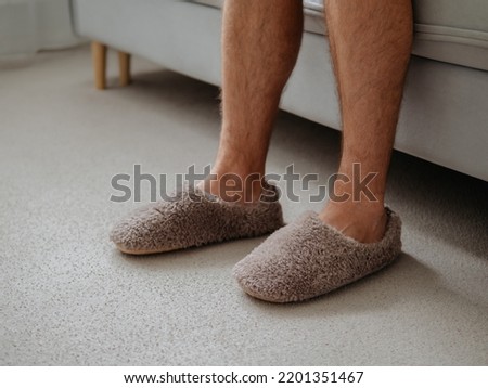 Cropped shot of unrecognizable man in soft plush warm slippers, sitting on sofa in morning time at cozy scandinavian home. Hairy legs of adult man in trendy warm slippers on floor
