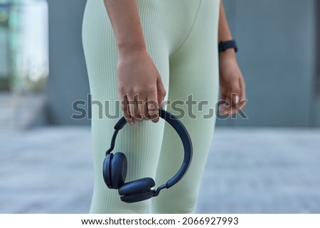 Cropped shot of unrecognizable person in leggings holds black stereo headphones uses modern technology for listening music during sport training outside. Female hands holding wireless acessory