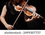 Cropped shot of unrecognizable professional female violinist in black dress playing classical music in dark studio with top light