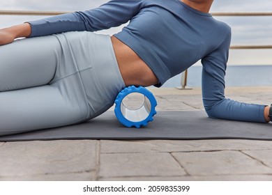 Cropped shot of unrecognizable slim woman uses foam roller for exercising stretches and massages muscles on fitness mat dressed in tracksuit tries to remove pain. Healthy lifestyle rehabilitation