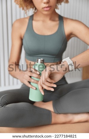 Cropped shot of unrecognizable sportswoman dressed in sportswear checks her progress on smartwatch sits crossed legs on floor rests after workout. Faceless sporty female involved in sport practice