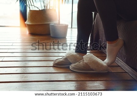 Cropped shot of unrecognizable woman putting on soft plush warm slippers, sitting on sofa in morning time at cozy home. Female wearing fluffy fur house shoes after waking up in bedroom in cold season