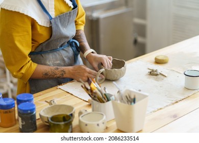 Cropped Shot Of Woman In Apron Shaping Pottery, Making Clay Mug During Ceramics Masterclass. Female Potter Standing At Wooden Table With Sculpting Tools And Art Supplies For Ceramist