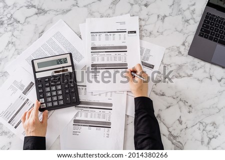 Cropped shot of woman calculating important monthly expenses, estimating total utility payments, using calculator and laptop, table with marble surface, writing with pen on paper