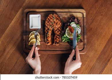 cropped shot of woman eating grilled salmon steak served on wooden board with lemon and lettuce