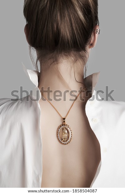 Cropped shot of a woman with golden necklace on her\
back. The oval pendant is made as Holy Virgin figure in open-work\
frame with crystals. The lady is dressed in white unbuttoned shirt\
back to front