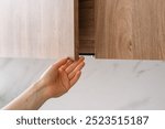 Cropped shot of woman hand is opening a wooden cabinet door in contemporary kitchen, highlighted by natural lighting. Female closed cupboard made with natural oak. Wood facade at home furniture