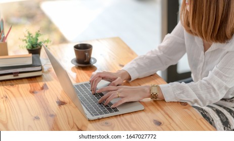 Cropped shot of a woman typing on laptop on wooden desk with coffee cup and office supplies while sitting next to window  - Shutterstock ID 1647027772
