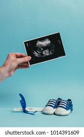 Cropped Shot Of Woman With Ultrasound Scan In Hand, Childish Shoes And Pregnancy Test With Ribbon Isolated On Blue