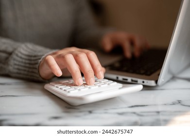 A cropped shot of a woman working at a table indoors, typing on a laptop keyboard, and using a calculator. multitasking skill, busy businesswoman, accounting - Powered by Shutterstock