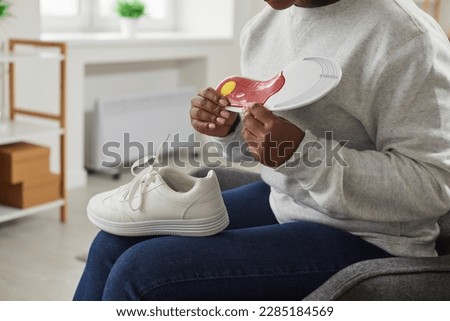 Cropped shot of a young African American woman with a sneaker on her lap sitting in an armchair at home and holding an orthotic arch support shoe insole in her hands. Footwear, feet health concept