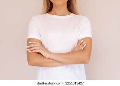 A cropped shot of a young blonde woman in a white t-shirt crossing her arms on her stomach isolated on a beige background. Closed pose