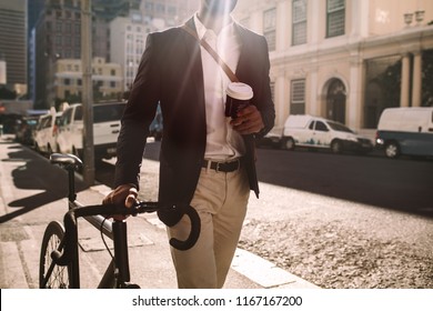 Cropped shot of young businessman going to work by bike. Man in suit holding a cup of coffee walking by the street with his bicycle.