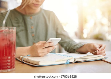 Cropped Shot Of Young Businesswoman Sitting At Cafe Looking At Her Organiser And In Calendar On Smart Phone Looking For Spare Day To Meet With Her Business Partners, Drinking Red Cold Cocktail