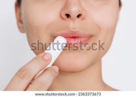 Cropped shot of a young caucasian attractive woman applying a hygienic lipstick on her lips on a light gray background. Moisturizing chapstick for dry lips