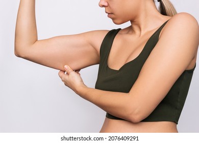 Cropped shot of a young caucasian blonde woman grabbing skin on her upper arm with excess fat isolated on a gray background. Pinching the loose and saggy muscles. Taking care of the body