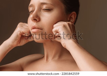 Cropped shot of young caucasian woman touching face with hands massaging her jaw isolated on a dark brown background. Rejuvenation, facelift, facefitness, skin care. Cosmetology and beauty concept
