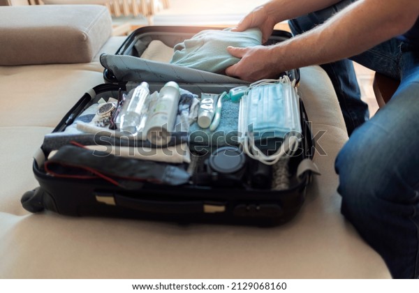 Cropped shot of a young men arranging clothes in\
suitcase for travel. Rear view of a man packing suitcase for\
business travel including face masks and airplane travel-sized\
antibacterial hand\
gels.