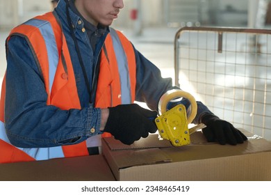 Cropped shot of young warehouse worker in safety vest and gloves packing metallic spare parts into boxes and sealing them with cellotape