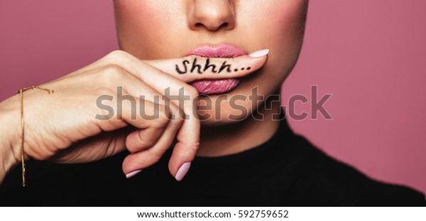 Cropped shot of young woman biting finger\
with shhh word. Young woman placing finger on lips asking shh\
against pink\
background.