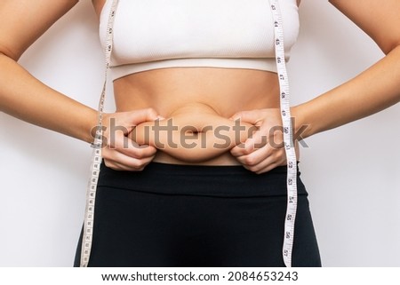 Cropped shot of a young woman 
with measuring tape around the neck holding herself by the fat on her stomach isolated on a white background.. Overweight, excess weight concept