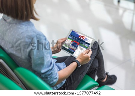 Cropped shot of a young woman sitting in the international airport terminal with a tablet in hands betting online at bookmaker's website. Gambling, money win concept. View from the back.