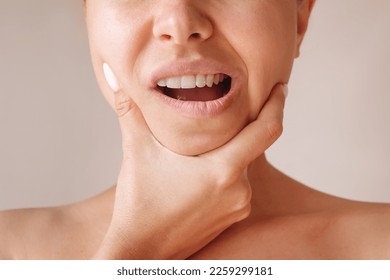 Cropped shot of a young woman suffering from jaw pain holding her chin isolated on a beige background. Inflammation of cervical lymph nodes, Diseases of ENT organs, facial, trigeminal nerve, toothache - Shutterstock ID 2259299181