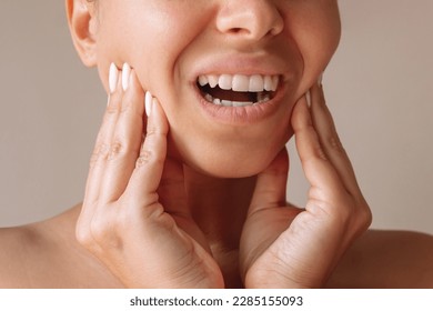 Cropped shot of a young woman suffering from jaw pain touching it with hands on a beige background. Inflammation of cervical lymph nodes, Diseases of ENT organs, facial, trigeminal nerve, toothache