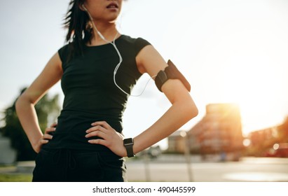 Cropped shot of young woman taking a break while on morning run. Fitness female with armband and earphones standing outdoors with her hands on hips. - Powered by Shutterstock