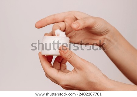 Cropped shot of a young woman taking moisturizing cream with her finger holding cosmetic jar in her hand isolated on a light  background. Skin care. A white thick smear of cream 