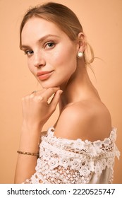 Cropped side portrait of a lady in a white lace blouse and a gold bracelet. The girl is wearing golden round stud earrings adorned with decorative white turquoise and posing on the peach background.   - Shutterstock ID 2220525747