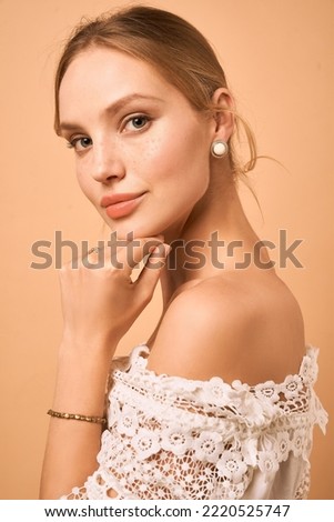 Cropped side portrait of a lady in a white lace blouse and a gold bracelet. The girl is wearing golden round stud earrings adorned with decorative white turquoise and posing on the peach background.  