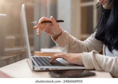 A cropped side view image of an Asian woman working on her laptop computer in a coffee shop, working remotely, pointing her finger and pen at laptop screen, typing on the laptop keyboard. - Powered by Shutterstock