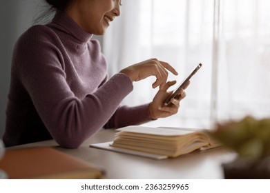 Cropped side view image of a beautiful and happy Asian woman using her smartphone at a table in her room. chatting, reading online blogs, responding to messages, scrolling on social media