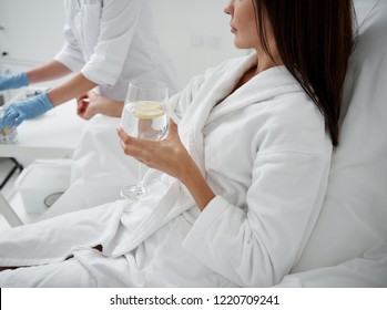 Cropped side view portrait of beautiful woman holding glass of lemon water while receiving intravenous treatment. Doctor in sterile gloves on background