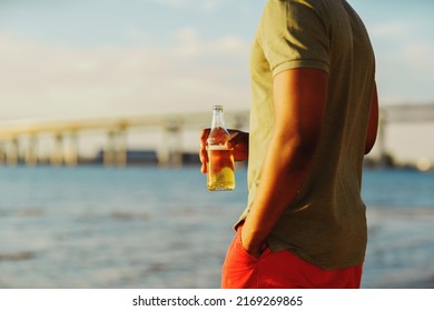 Cropped Side View Of Unrecognizable Young Black Man Drinking Beer From Bottle And Looking At Sea View Standing On Beach On Summer Day, His Hand In Pocket