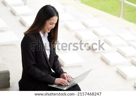 Cropped sideview of an Asian business woman In a black suit and black skirt working on her laptop, being focused on work at outside the office building. Business and financial  accounting concept.