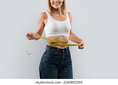 Cropped of slim blonde woman in white top and dark jeans measuring her waist with measure tape, isolated on grey studio background, copy space. Weight loss, slimming concept - Powered by Shutterstock