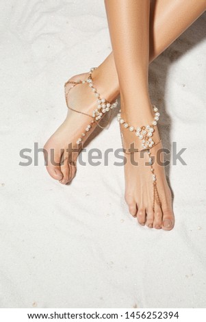 Cropped top view shot of lady's crossed legs on the sandy background. The girl with light brown pedicure is wearing a golden anklet adorned with chains and pearls. Trendy women's summer accessory.