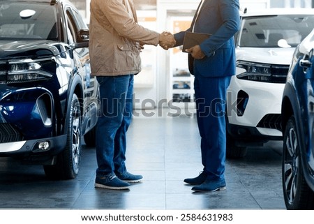 Cropped of two men are standing inside a bright and spacious car showroom. They are shaking hands with each other, appearing satisfied and confident after finalizing a deal on a new vehicle.