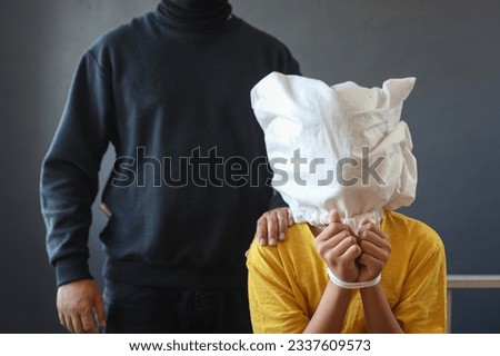 Cropped unrecognizable man maniac in black outfit standing behind kidnapped little boy with paper bag over his head and tied up. Child abuse, kidnap and trafficking crime concept