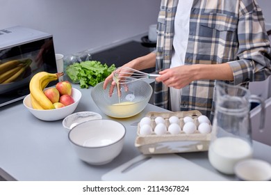 Cropped unrecognizable young woman kneading dough with whisk for banana pancakes in bowl in kitchen with white, light interior. Caucasian female in casual checkered shirt at home alone