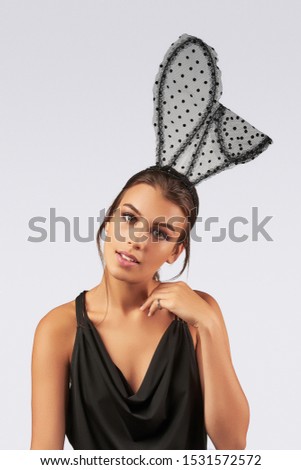 Cropped upper body's shot of a dark-haired girl, posing on a grey background. She is wearing black v-neck tank top and black headband with polka dot mesh rabbit ears on her head. 