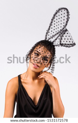 Cropped upper body's shot of a dark-haired girl, posing on a grey background. She is wearing black v-neck tank top and black headband with polka dot mesh rabbit ears and black polka dot mesh veil.